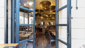 An interior with open doors leading to a dining area with wooden tables and chairs, a bar area with stools, and hanging pendant lights. - Saifi Suites Hotel (Beirut)