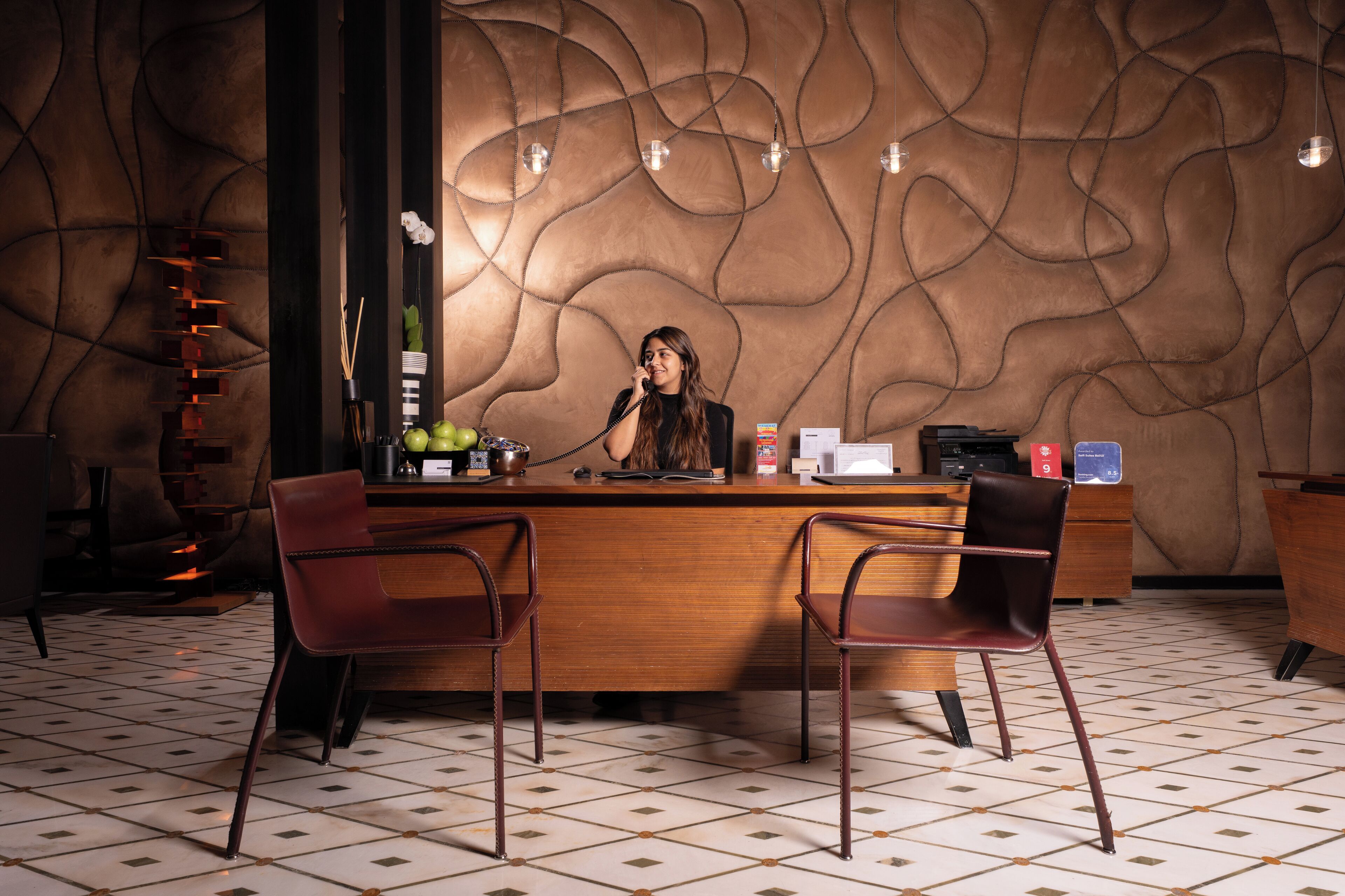 A reception area with a wooden counter, two brown chairs, a decorative wall, and hanging lights.