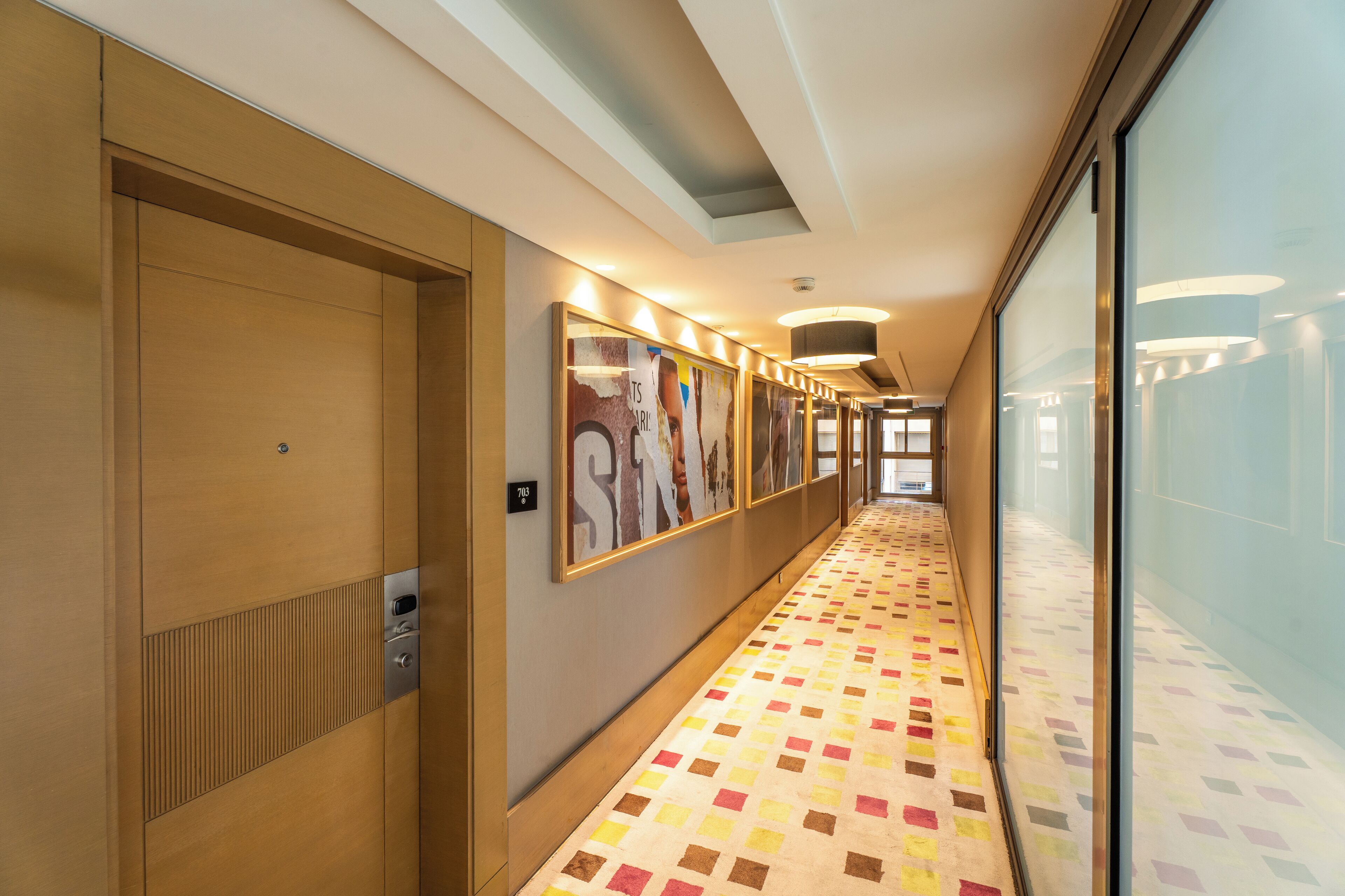A hotel corridor with wooden doors, framed artwork on the walls, and a patterned floor.