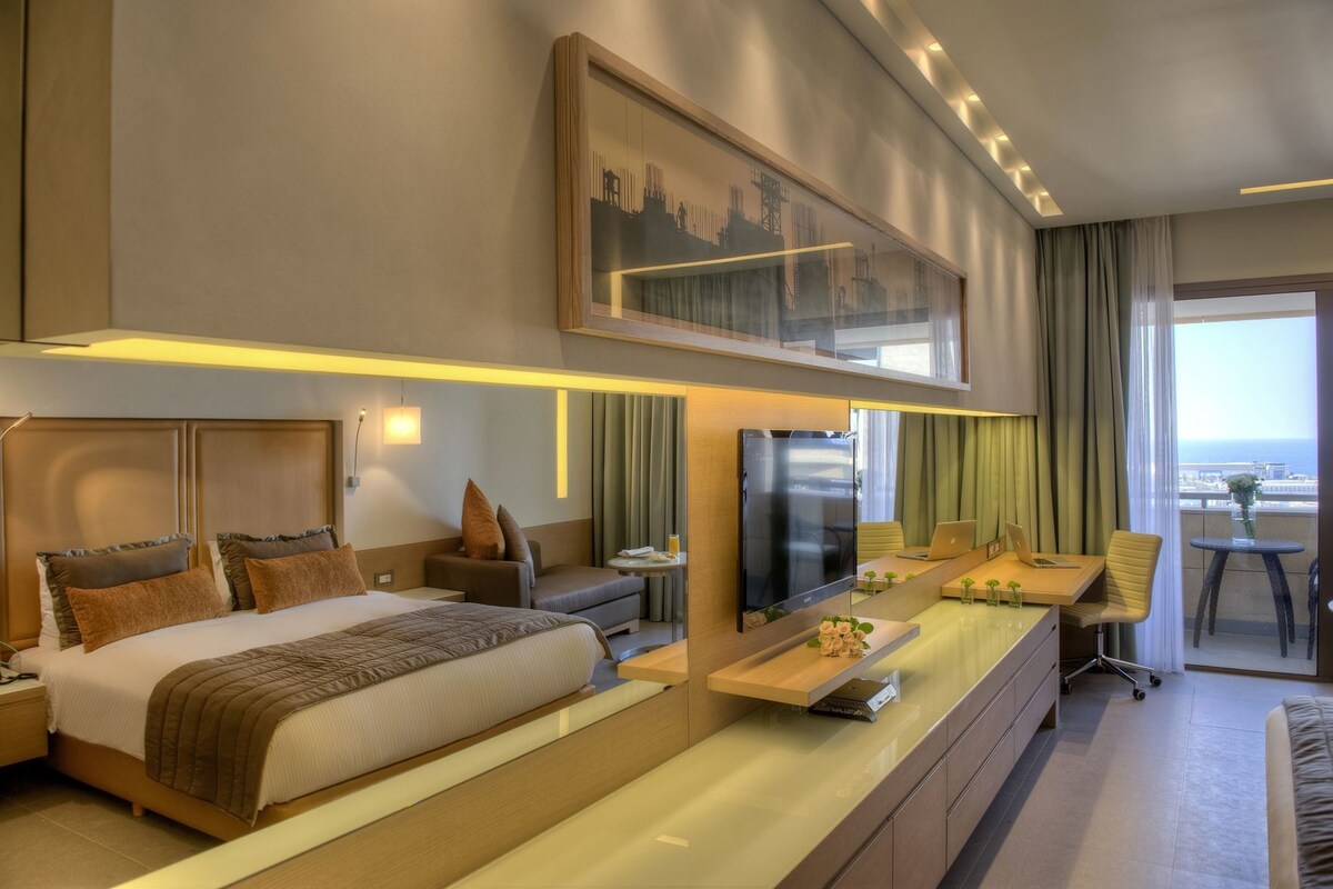 Ein modernes Hotelzimmer mit einem großen Bett, einem Schreibtisch mit einem Laptop, einem Flachbildfernseher und einem Balkon mit Aussicht.