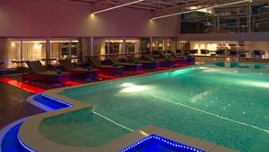 Piscina interior com teto de vidro, cadeiras de descanso e área ao redor da piscina com iluminação LED azul.