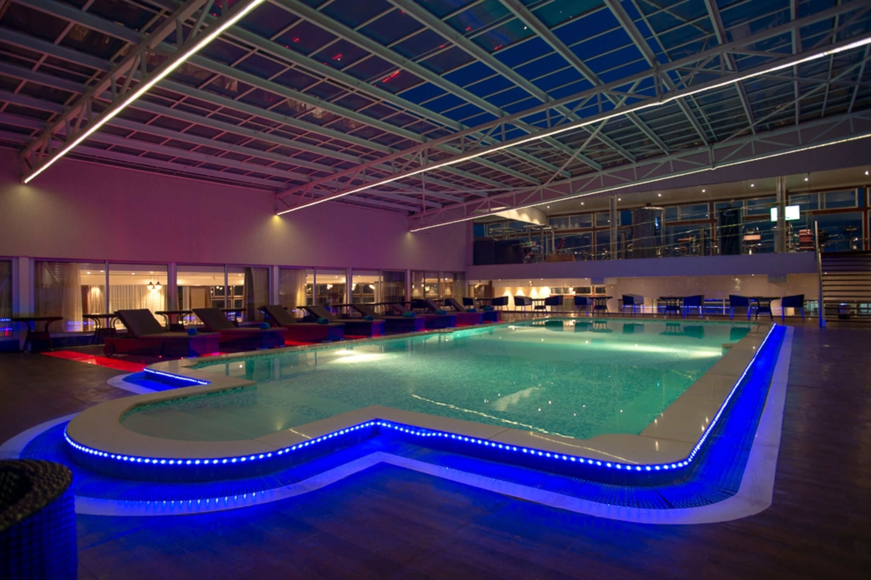 An indoor swimming pool with a glass ceiling, lounge chairs, and a poolside area with a view of the city.