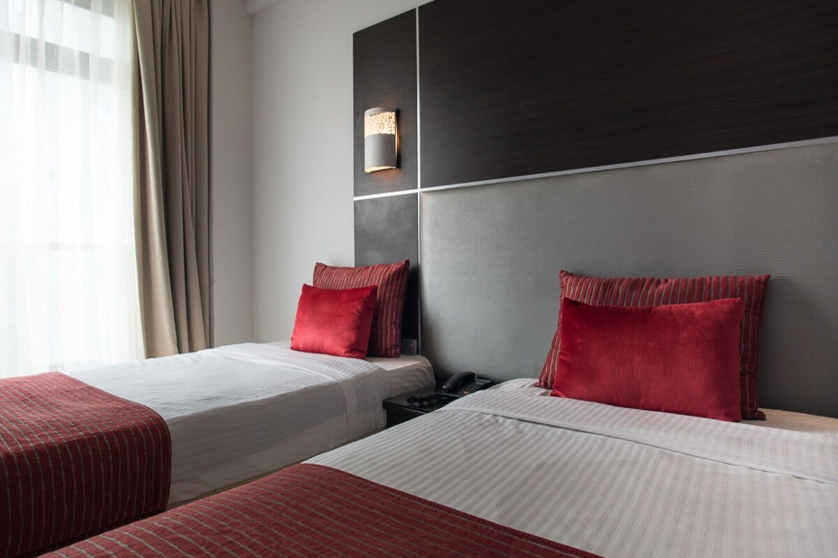 A hotel room with two beds, each with red pillows and white bedding. There is a bedside table with a phone and a wall-mounted light fixture.
