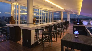 Uma área de bar moderna com pé direito alto, grandes janelas e vista para a cidade.