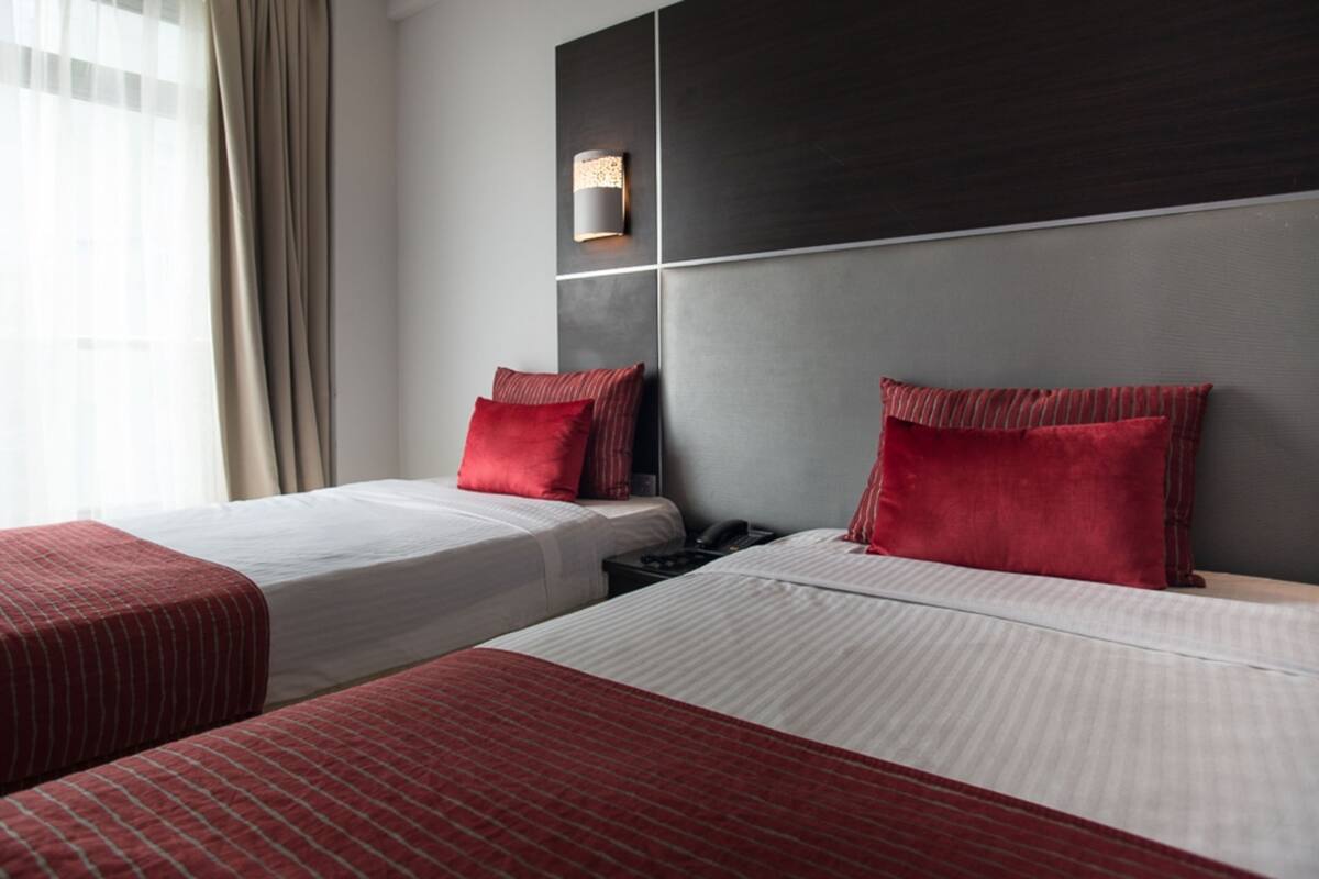 A hotel room with two beds, each with red and white bedding, a bedside table with a phone, and a wall-mounted light fixture.