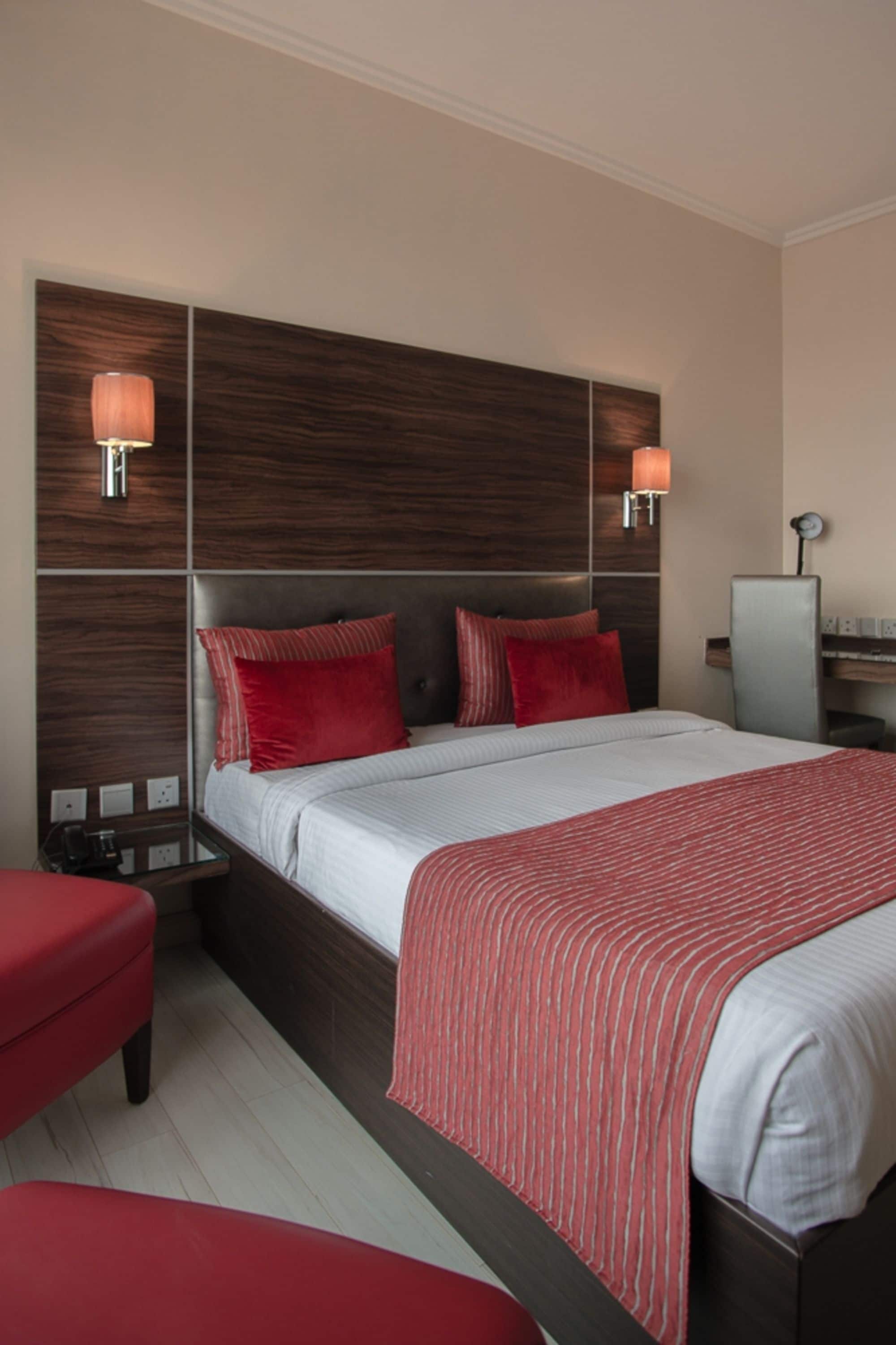 A hotel room with a large bed, red pillows, a red upholstered chair, and a desk with a lamp.