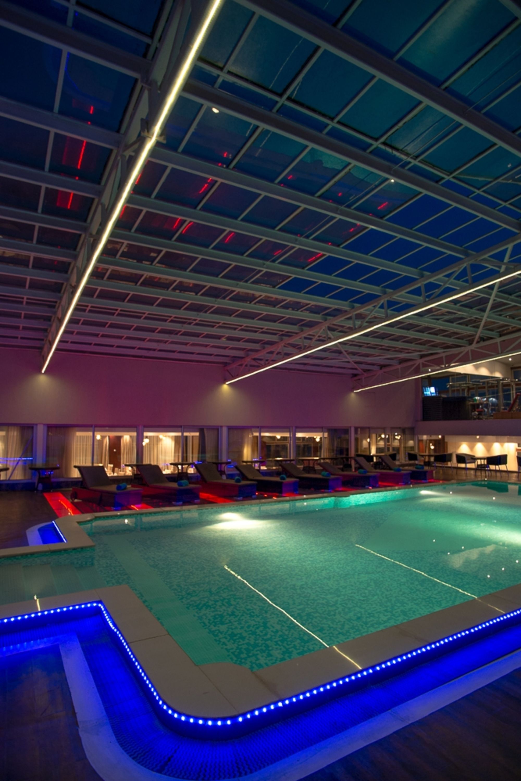 An indoor swimming pool with a glass ceiling, lounge chairs, and a bar area.