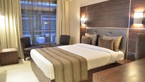 Quarto de hotel moderno com uma cama grande, uma mesa de escritório e uma cadeira. Há uma janela com cortinas, uma luminária na parede e vista para a cidade.