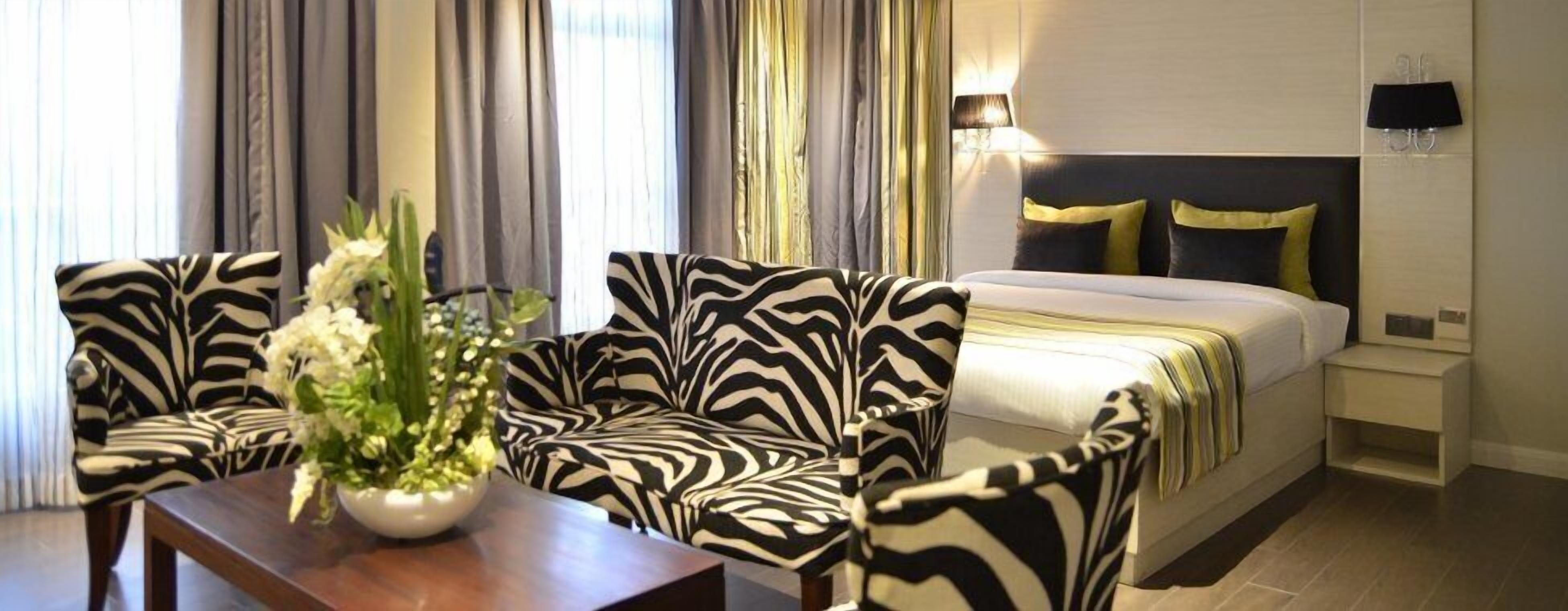 A hotel room with a zebra-patterned sofa set, a bed with a gold and black headboard, and a bedside table with a lamp.