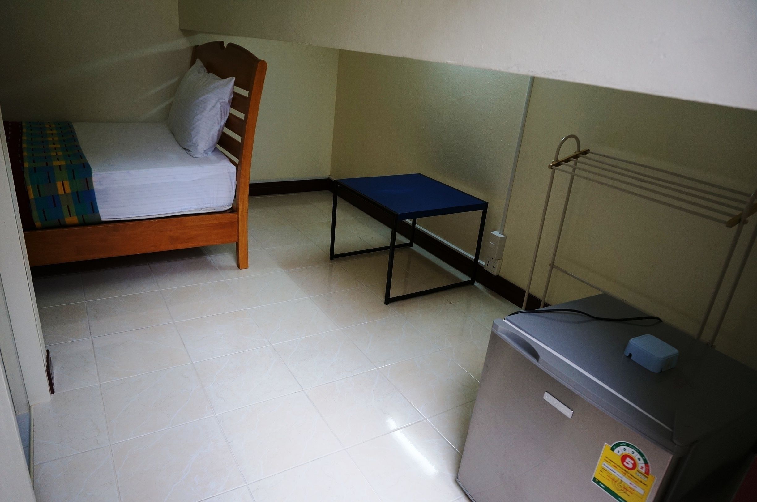 Una habitación pequeña y funcional con una cama individual, una mesita y un refrigerador compacto.