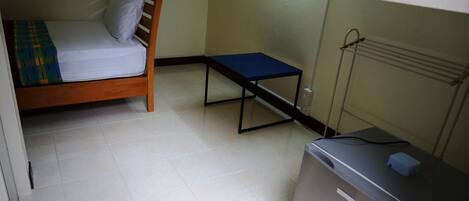 Una habitación pequeña y funcional con una cama individual, una mesita y un refrigerador compacto.