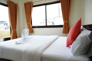 A neatly made bed with white bedding and a red pillow, curtains, and a window with a view of buildings.