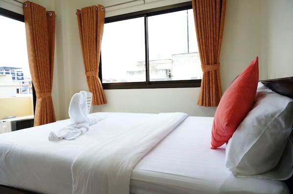 Una cama bien hecha con ropa blanca y una almohada roja, cortinas y una ventana con vistas a edificios.