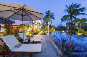 Zona de piscina con sillones, una sombrilla grande y una piscina iluminada con palmeras.
