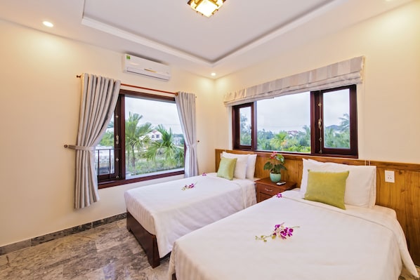 A hotel room with two beds, a window with curtains, an air conditioning unit, and a view of greenery outside.