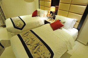 Habitación de hotel con dos camas, cada una con ropa de cama blanca y almohadas rojas, una mesita de noche con lámpara y un cabecero con diseño acolchado.