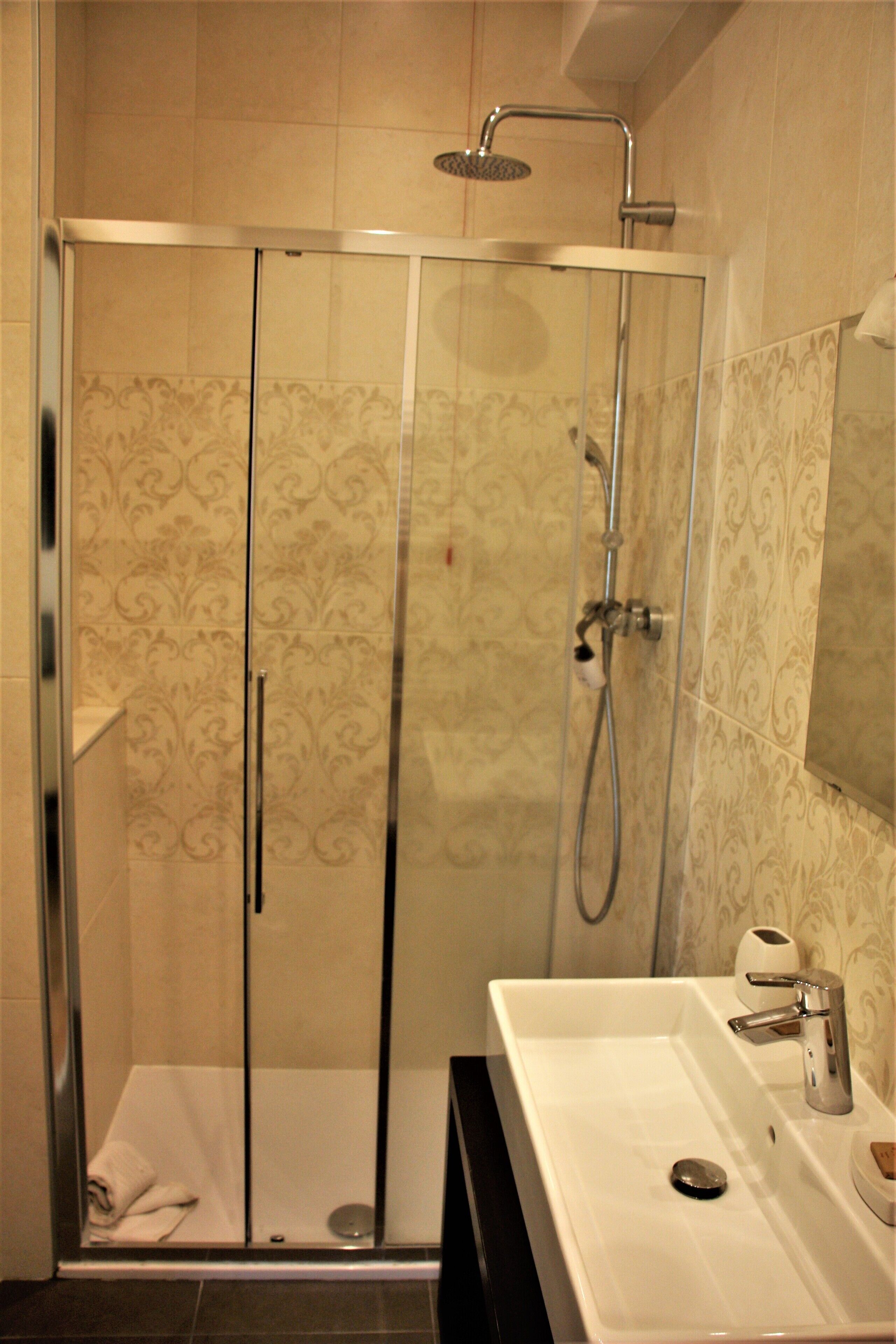 A glass-enclosed shower with a handheld showerhead, a white sink with a soap dispenser, and a mirror above the sink.