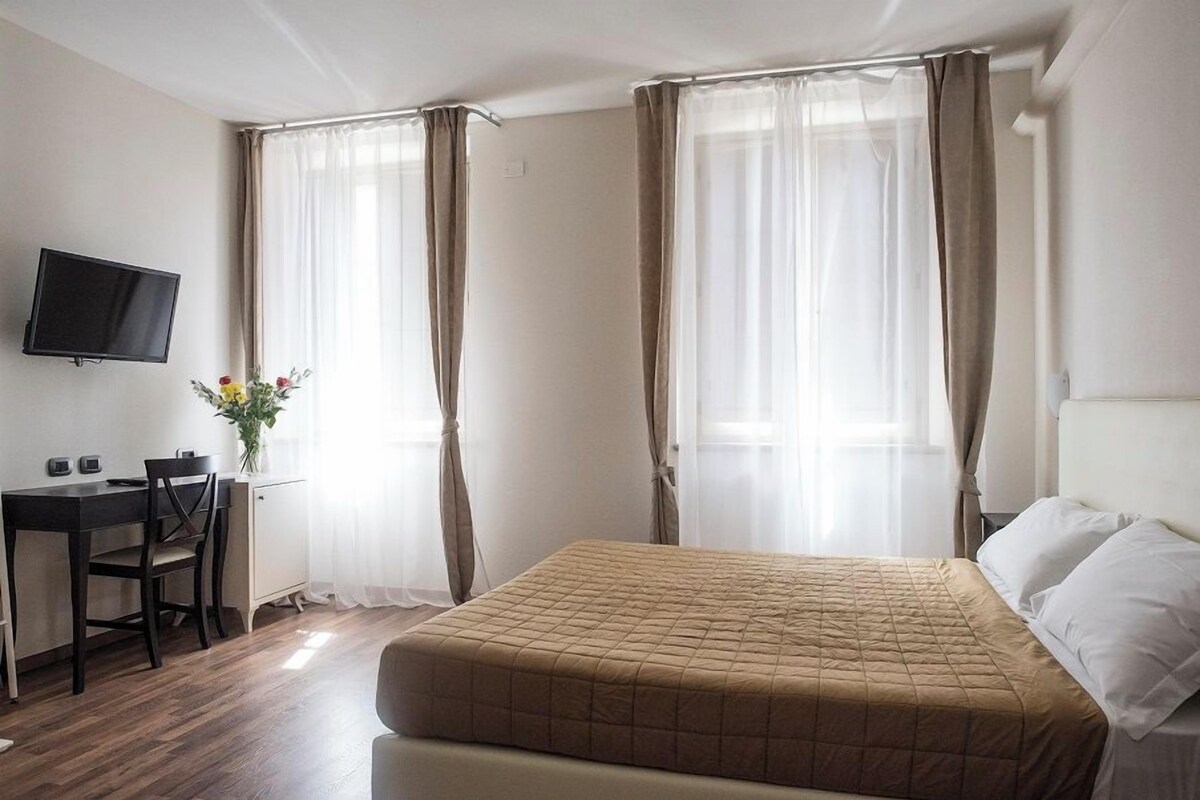 A hotel room with a bed, a desk, a chair, a TV, and a window with curtains.