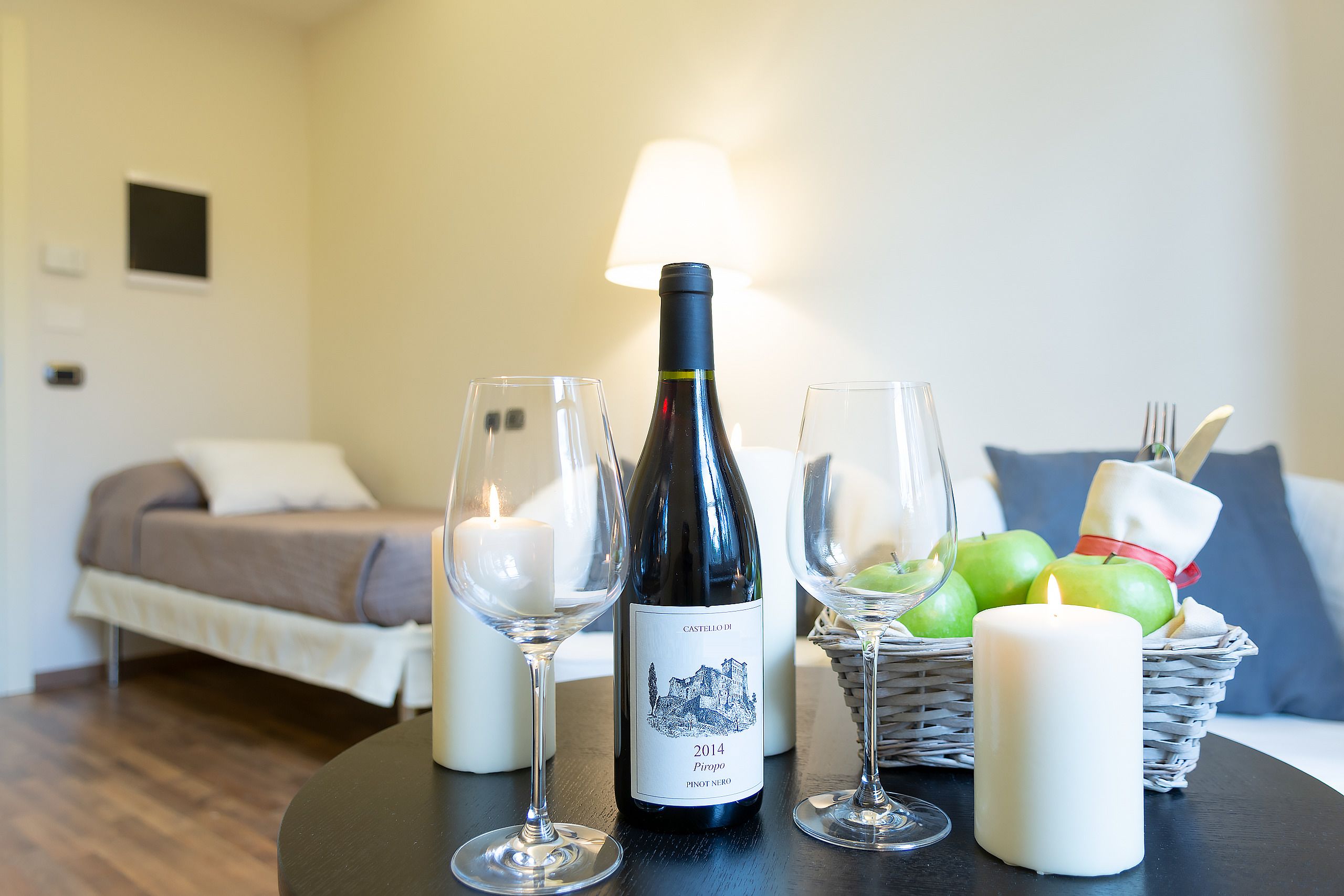 A room with a bed, a table with a bottle of wine, glasses, a candle, and a basket of apples.