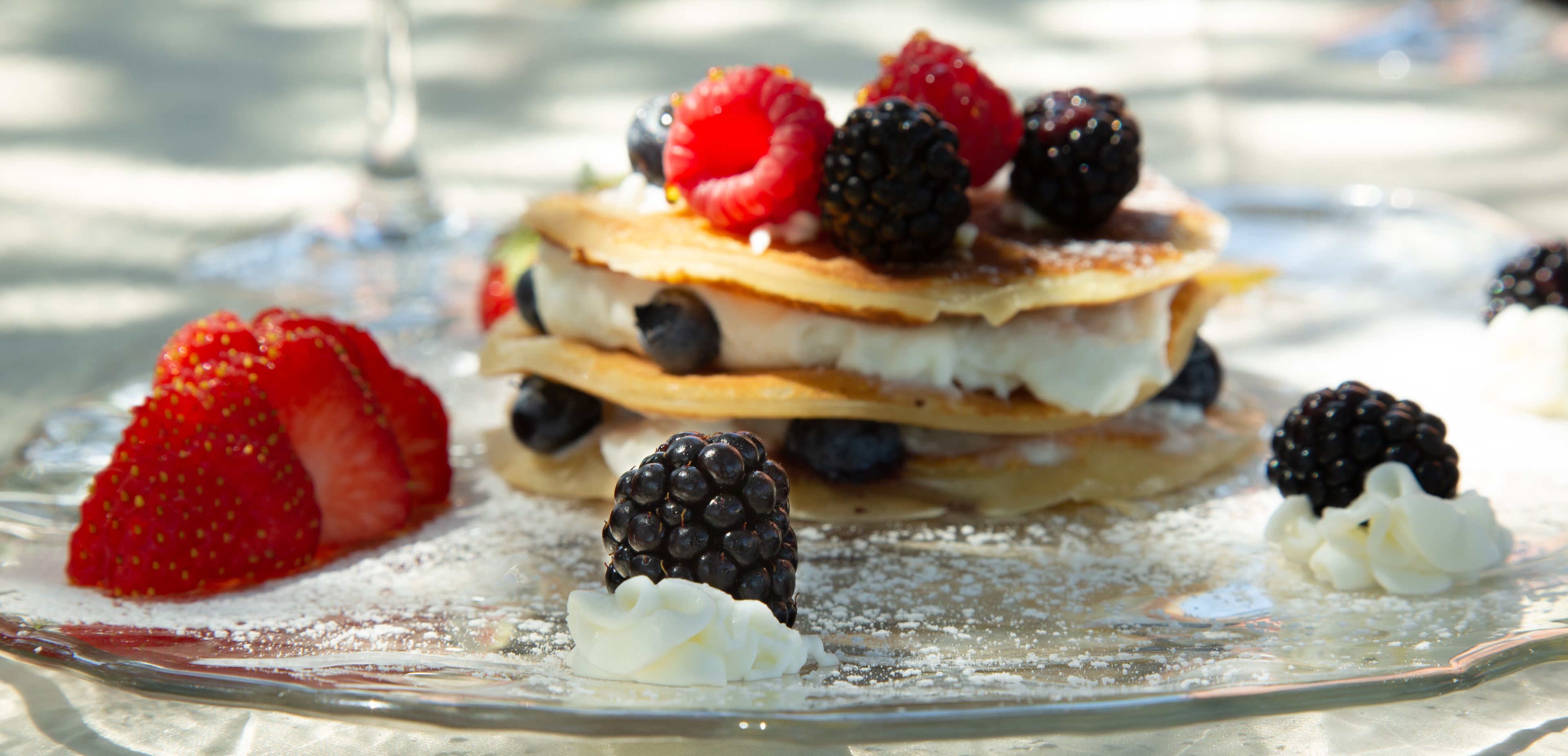 Una pila di pancake con frutti di bosco e panna montata su un piatto di vetro.