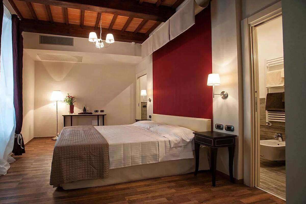 A bedroom with a bed, bedside tables, a nightstand, a red accent wall, a wooden ceiling, and a window with curtains.