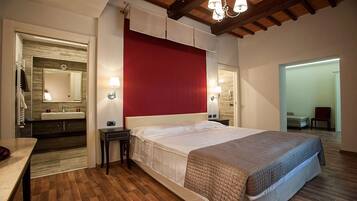 A bedroom with a large bed, a bedside table, a nightstand, a red accent wall, a mirror, and a bathroom visible through an open door.