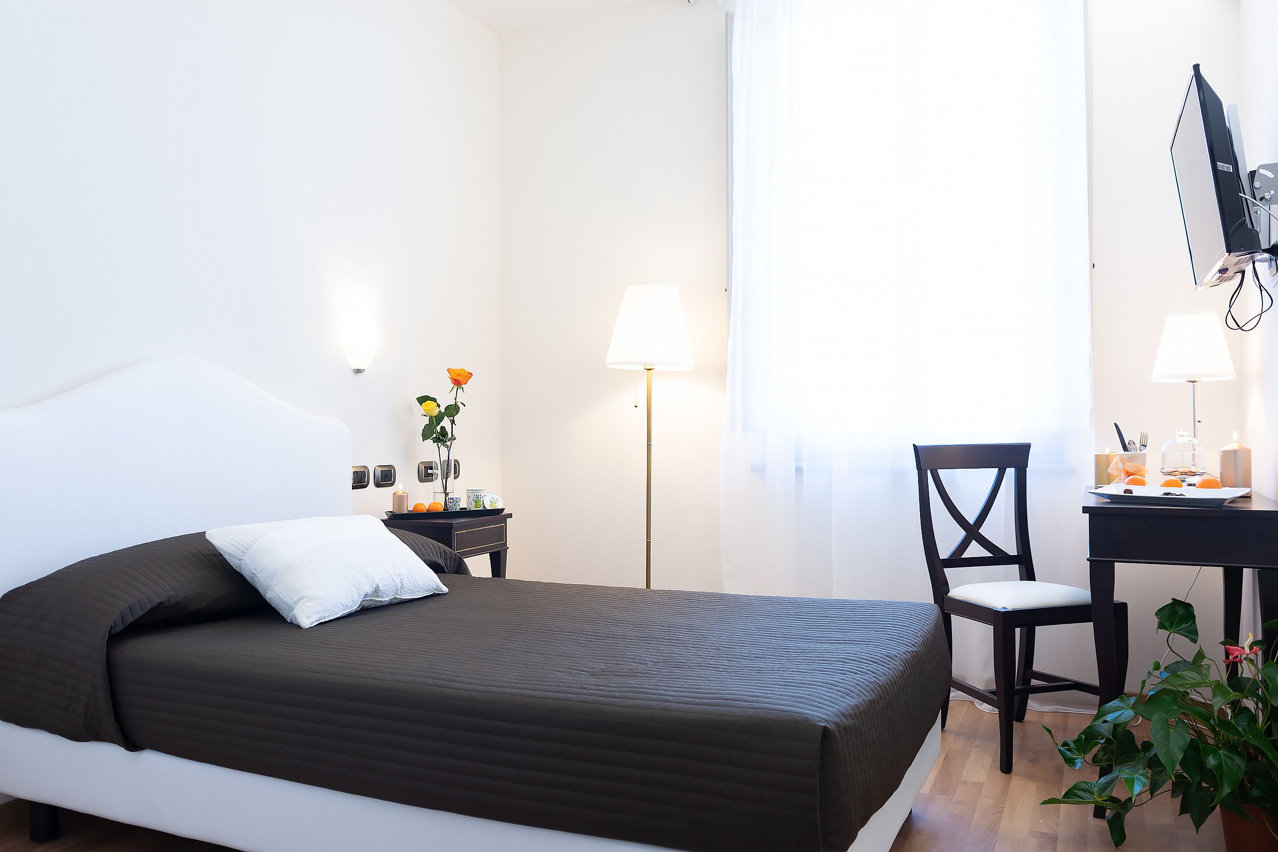 A neatly arranged bedroom with a bed, bedside table, lamp, chair, desk, and a potted plant.