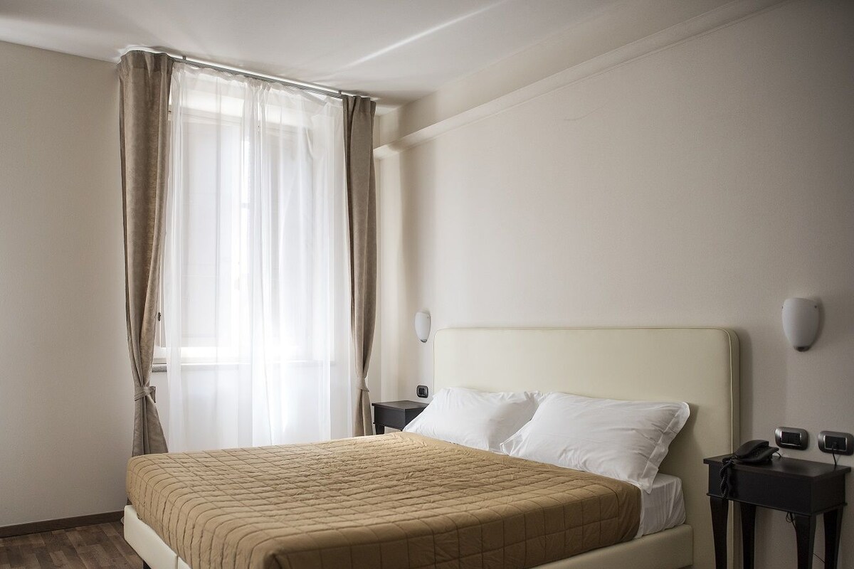 Una habitación de hotel con cama, mesita de noche, lámpara de pared y ventana con cortinas.