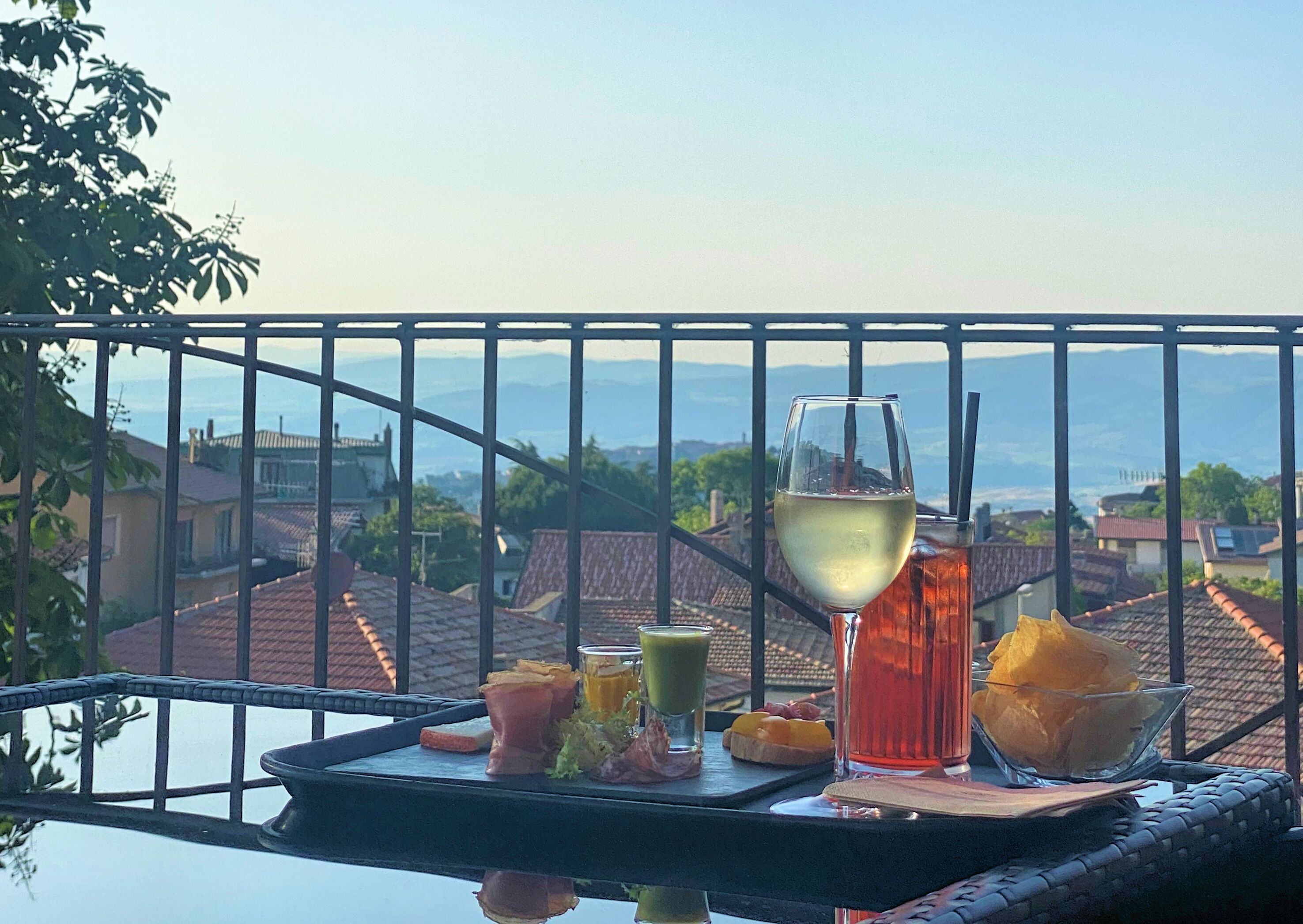 Uma varanda com uma mesa posta para refeição, incluindo um copo de vinho branco, um coquetel e diversos petiscos.