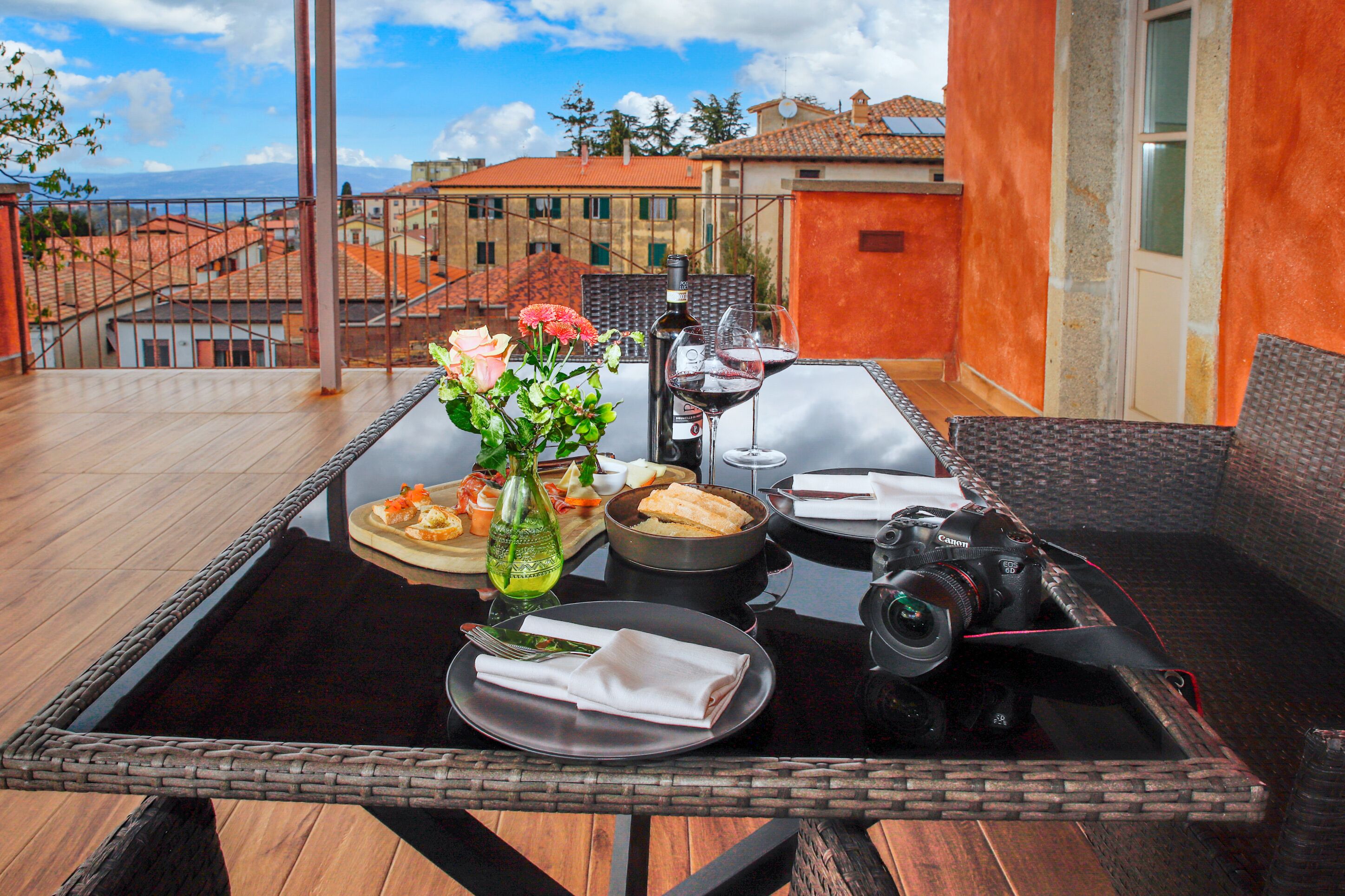 A dining table with a meal, a camera, and a bottle of wine on a balcony overlooking buildings.