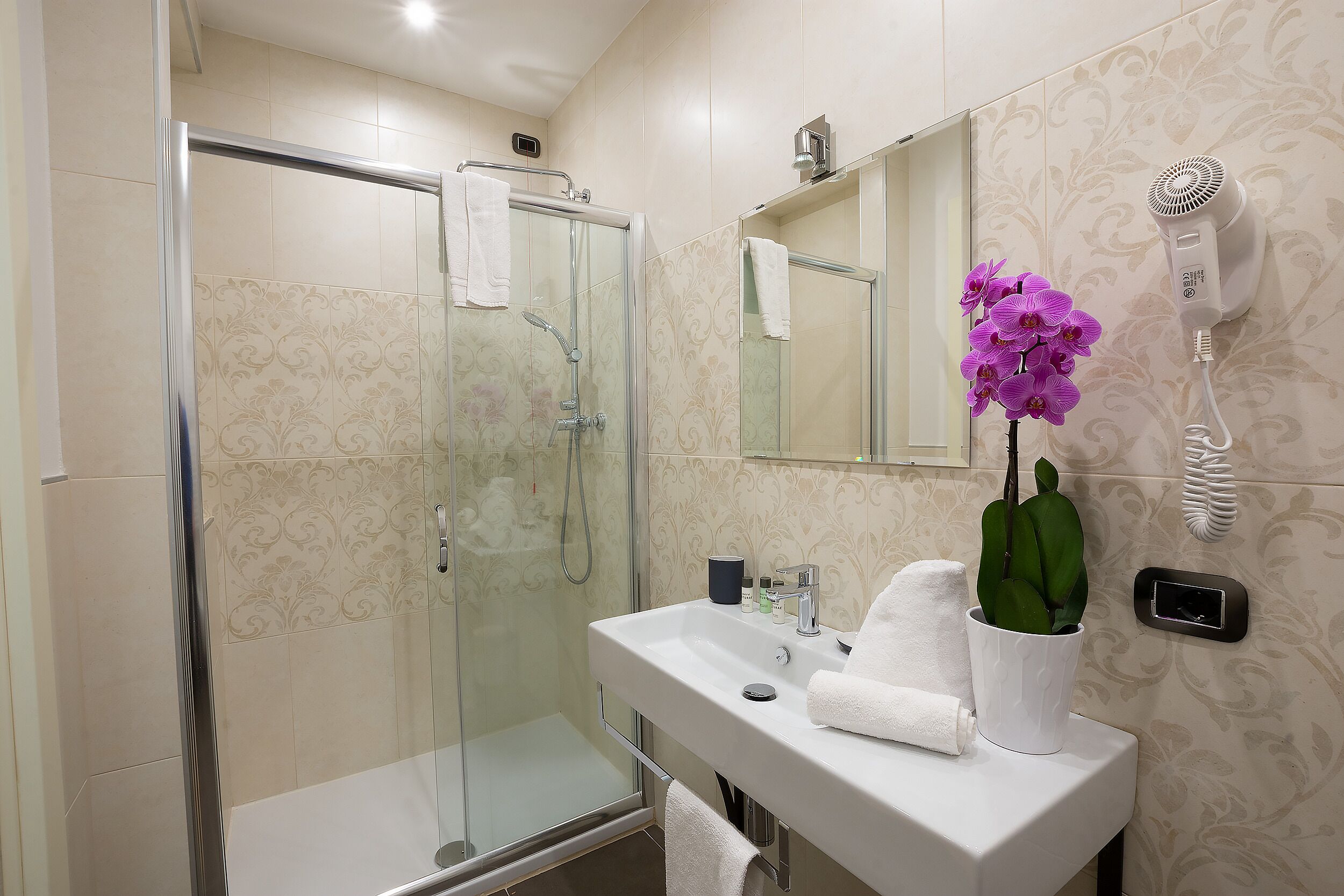 Un baño con cabina de ducha de vidrio, un lavamanos blanco con un espejo encima y un secador de pelo montado en la pared.