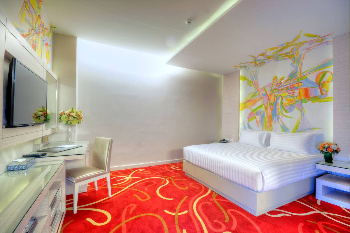 Une chambre d’hôtel moderne, dotée d’un grand lit, d’un bureau avec une chaise, d’une télévision et d’une peinture abstraite aux couleurs vives accrochée au mur.