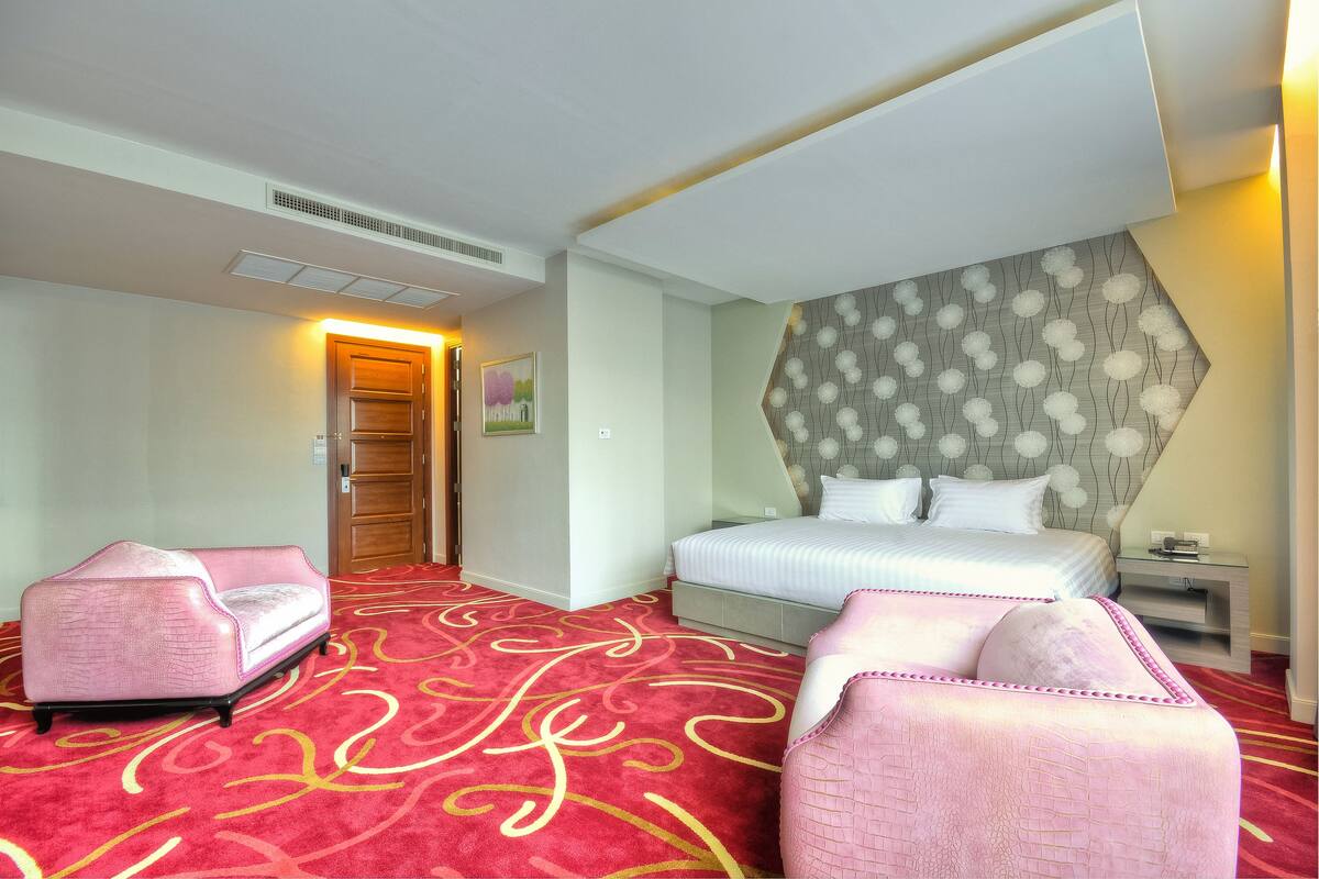 Une chambre d’hôtel avec un grand lit, deux fauteuils roses, une moquette rouge à motifs et un panneau décoratif mural.