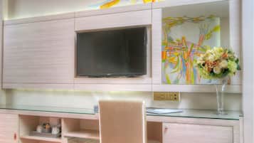 A modern hotel room with a large flat-screen TV, a glass desk, a white chair, and a vibrant red carpet.