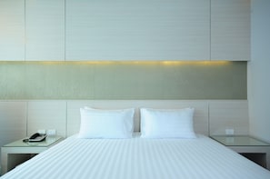A hotel room with a bed, two white pillows, a glass bedside table with a telephone, and a wall-mounted light.