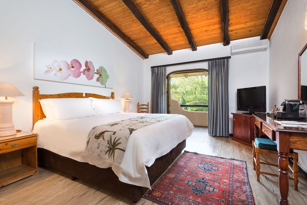 Un dormitorio con techo de madera, una cama con colcha estampada, un escritorio con televisor y vista a zonas verdes.