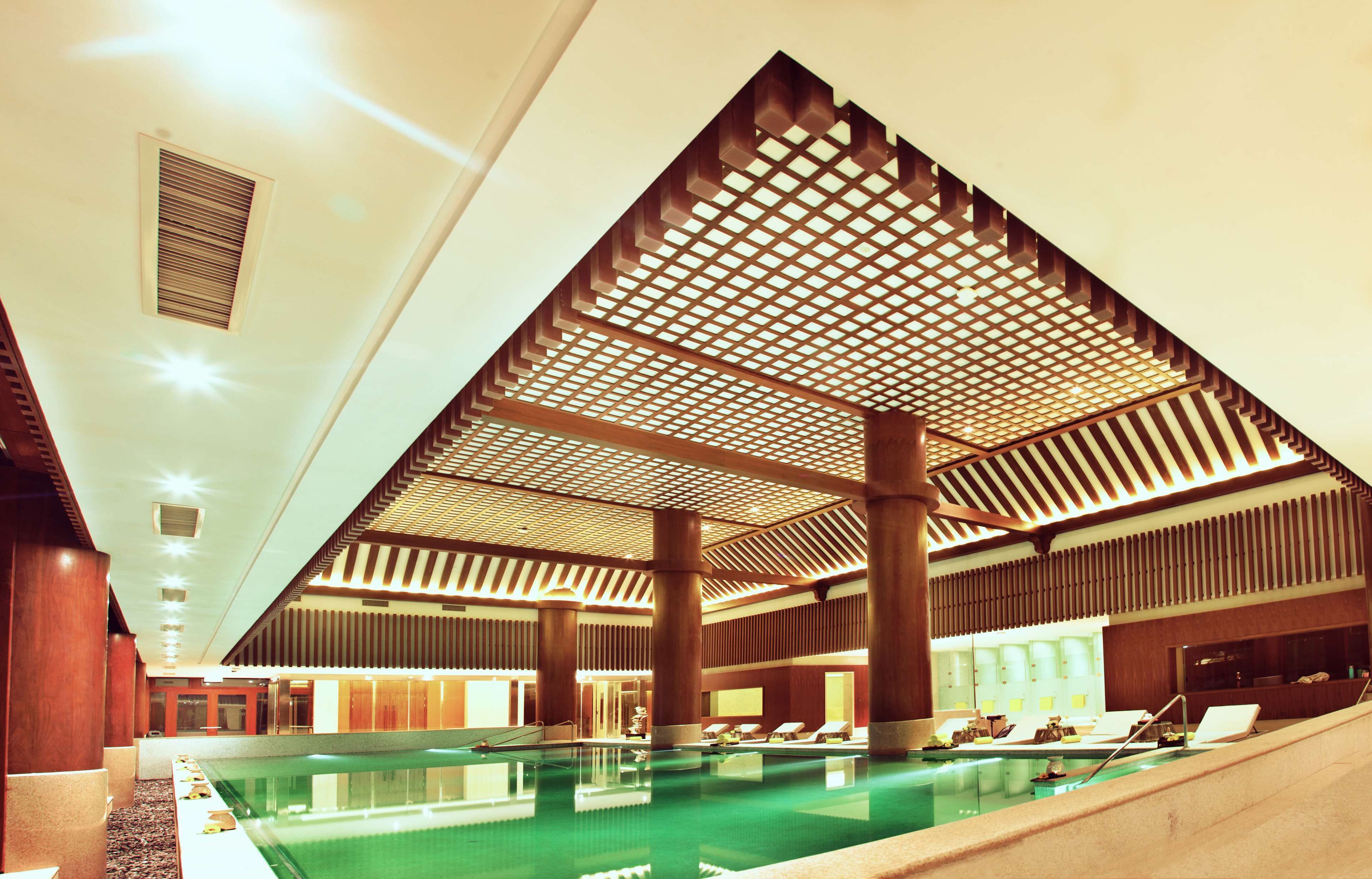 An indoor swimming pool with a geometric ceiling structure, lounge chairs, and a reception area.