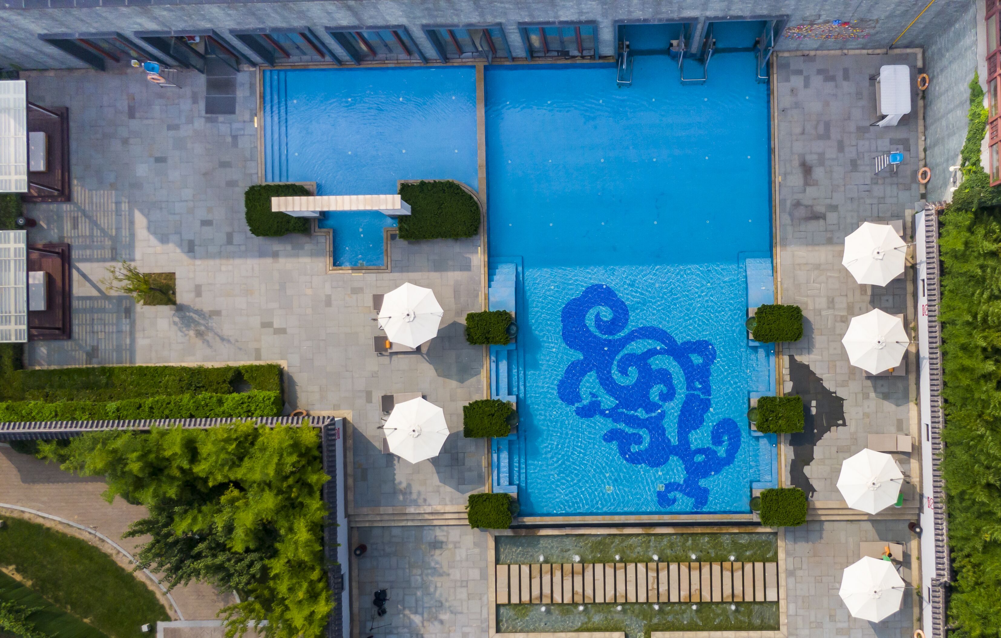 An aerial view of a hotel with two large swimming pools, surrounded by outdoor seating areas and greenery.