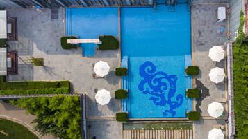An aerial view of a hotel with two large swimming pools, surrounded by outdoor seating areas and greenery.