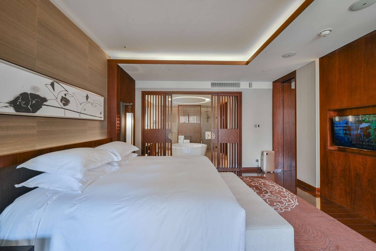 A modern hotel room with a large bed, a television, a bathroom with a bathtub, and a wooden floor.