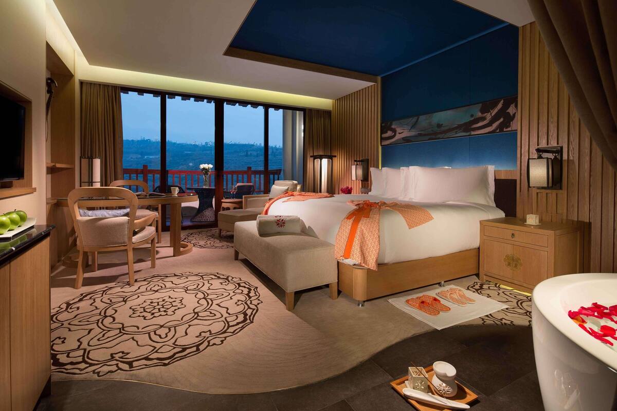 A hotel room with a large bed, a desk, a chair, a sofa, a balcony with a view, and a bathtub with rose petals.
