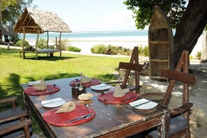 Un comedor al aire libre con mesa de madera, individuales rojos y sillas, con vista a una playa y una cabaña con techo de paja al fondo.