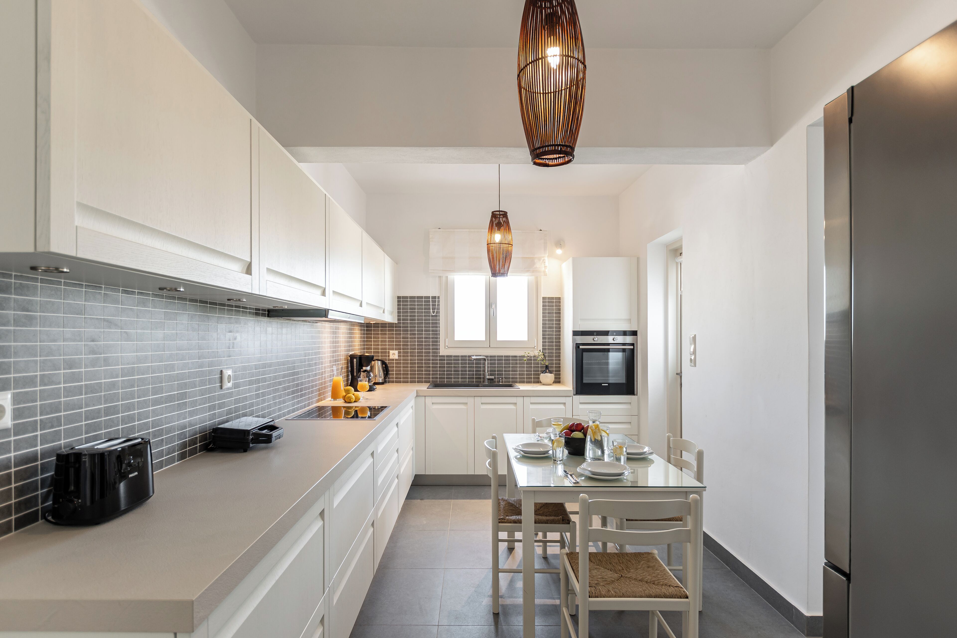A modern kitchen with white cabinetry, a grey tiled backsplash, a stainless steel refrigerator, and a dining area with a table and chairs.