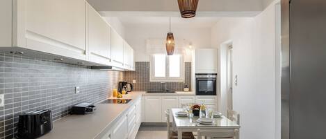 A modern kitchen with white cabinetry, a grey tiled backsplash, a stainless steel refrigerator, and a dining area with a table and chairs.