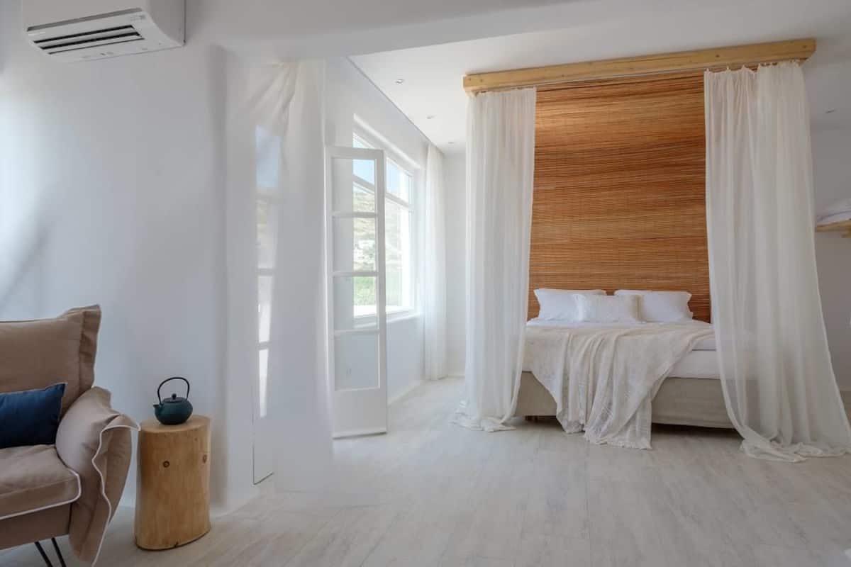 Un dormitorio con cama, cabecera de madera, cortinas blancas y vistas por una ventana.