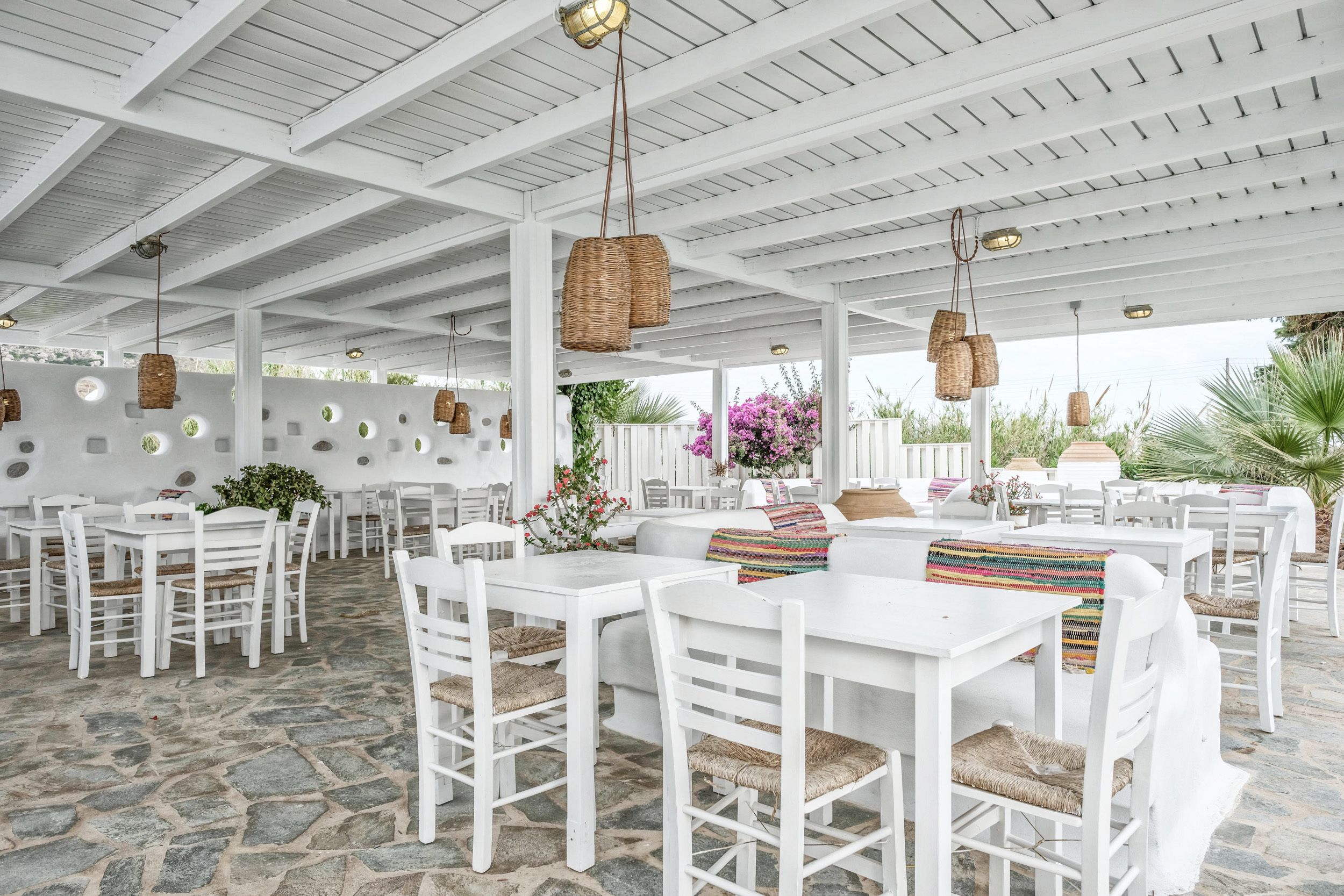 An outdoor dining area with white tables and chairs, stone flooring, and hanging wicker lamps.