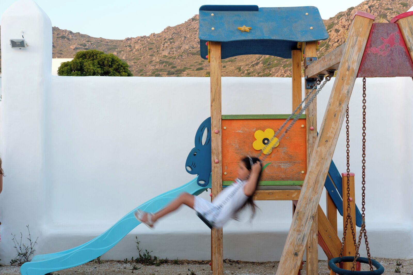 A child on a playground swing set with a slide, set against a white wall and a mountainous backdrop.