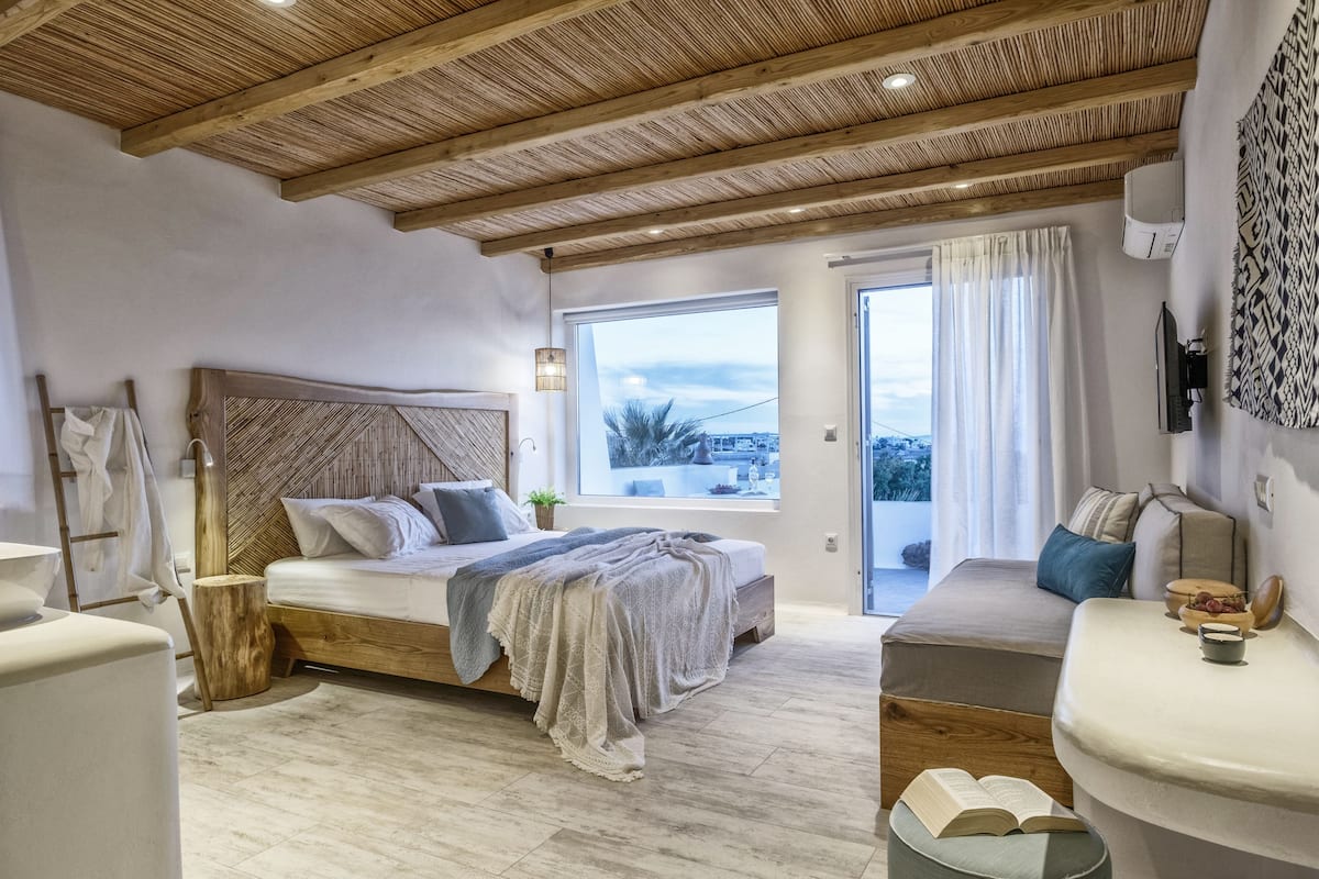 Un dormitorio con techo de madera, un ventanal con vistas, una cama con cabecera y una mesita de noche con libros.