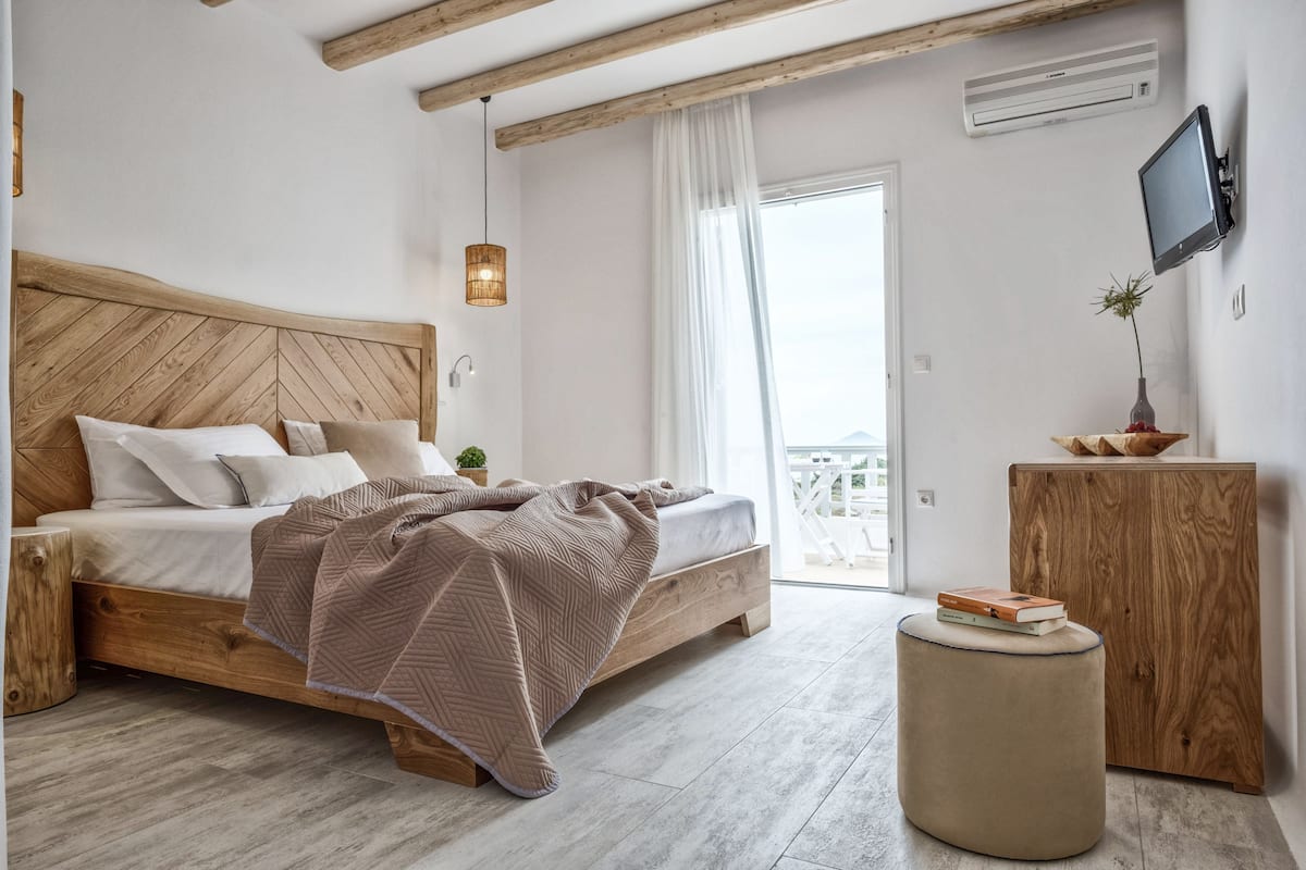 Un dormitorio con una cama de madera, una mesita de noche, un otoman redondo, un armario de madera y un televisor instalado en la pared.