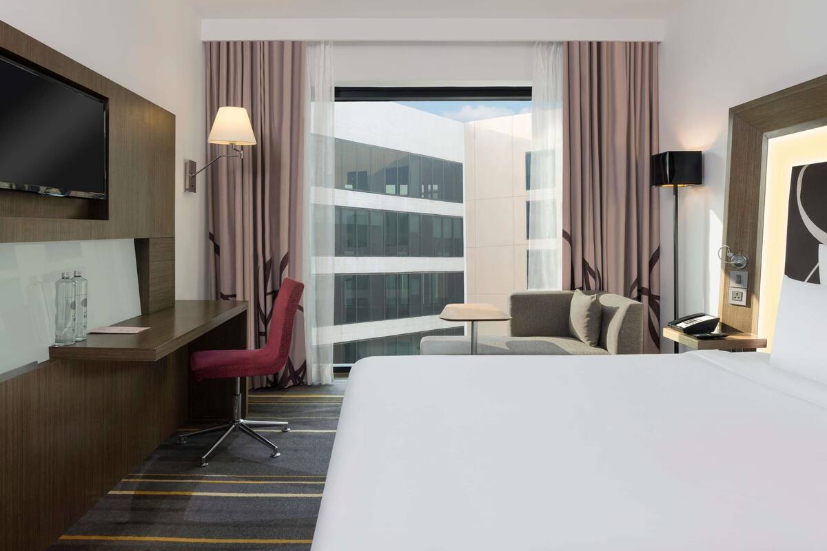 Hotelkamer met een bed, bureau, stoel, televisie, lamp en grote ramen met gordijnen.